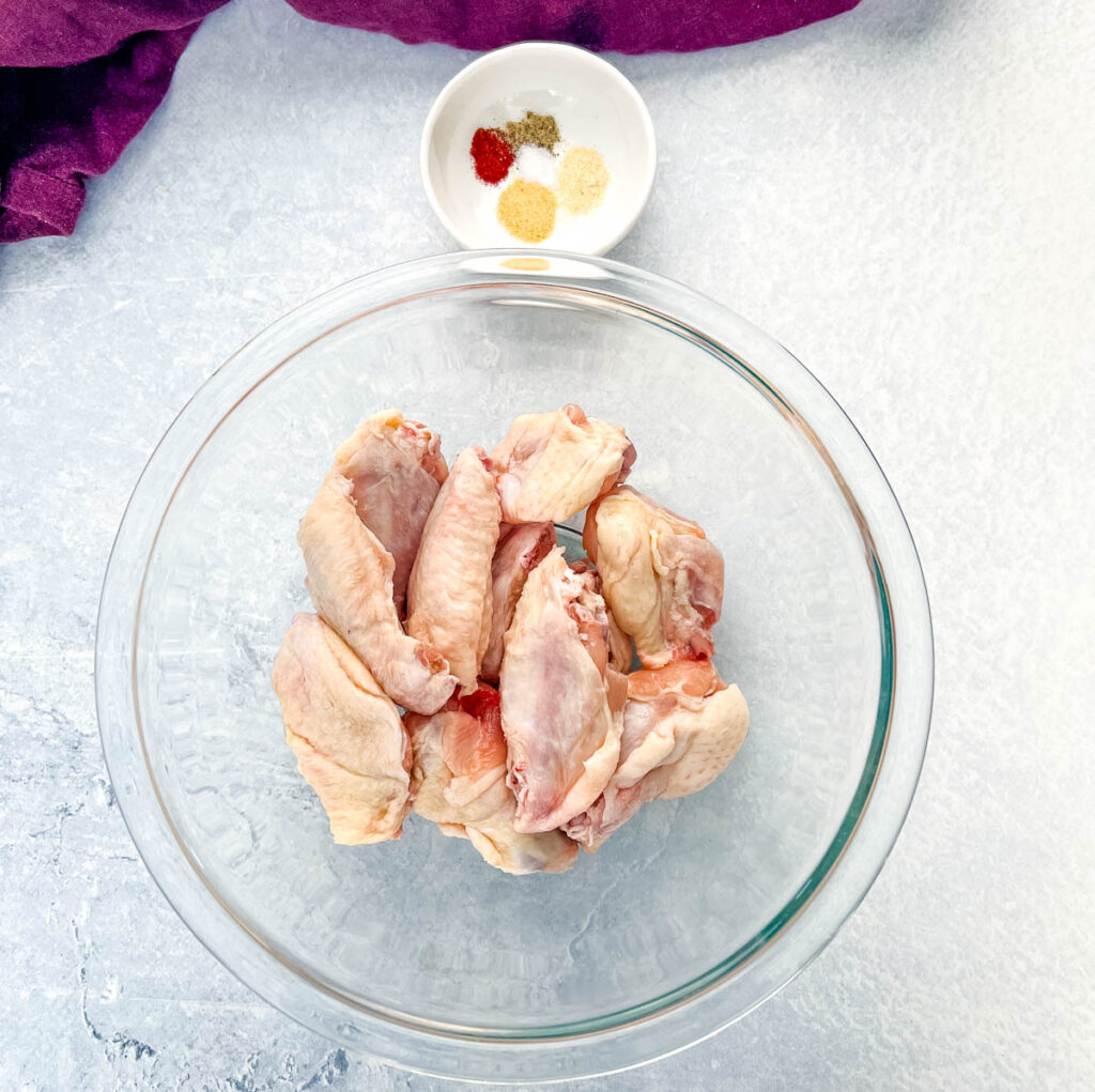raw chicken wings in a glass bowl along with a bowl of spices