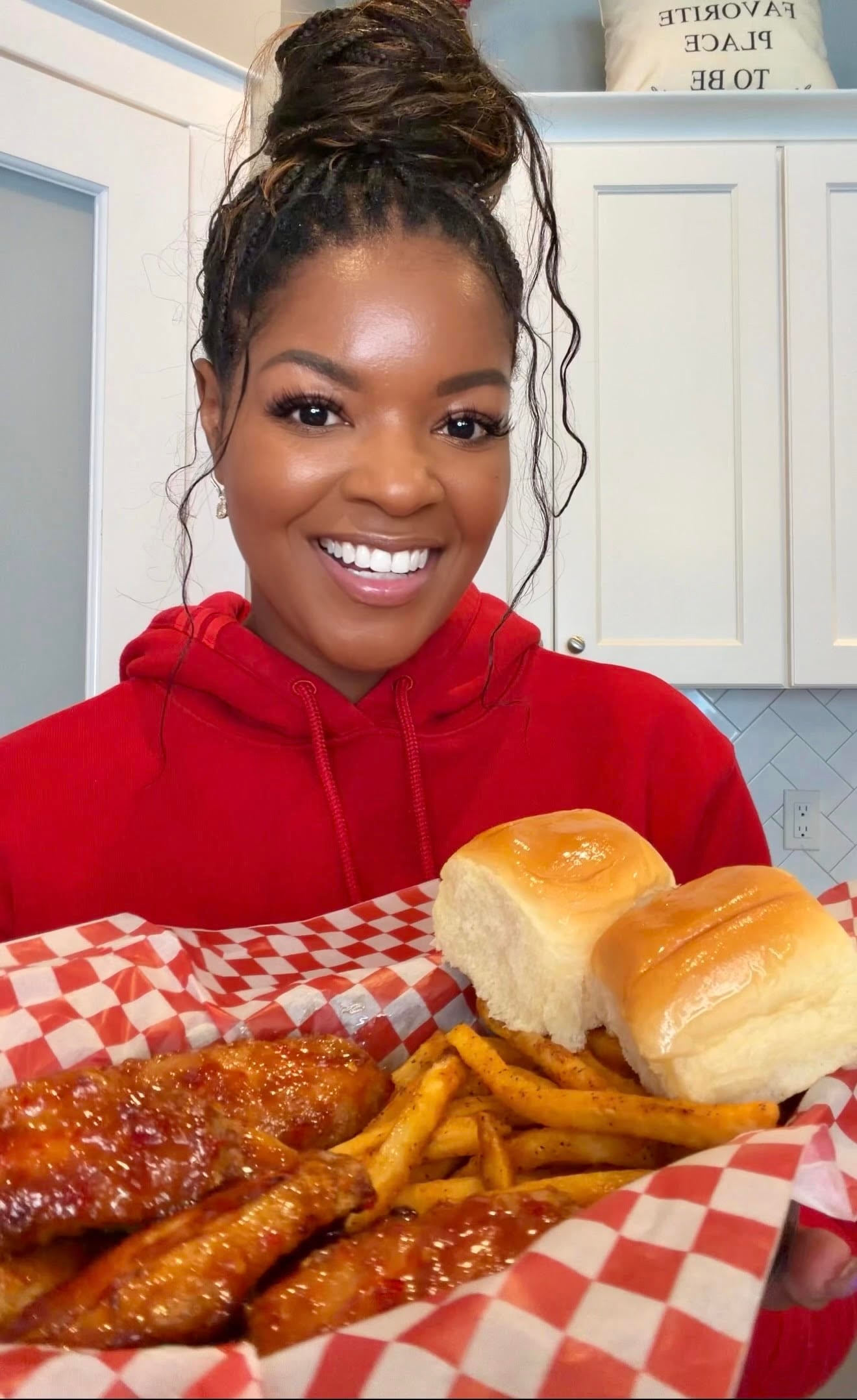 Smiling woman in a red hoodie holding a basket of crispy sweet chili chicken wings, seasoned fries, and buttery dinner rolls in a bright kitchen.