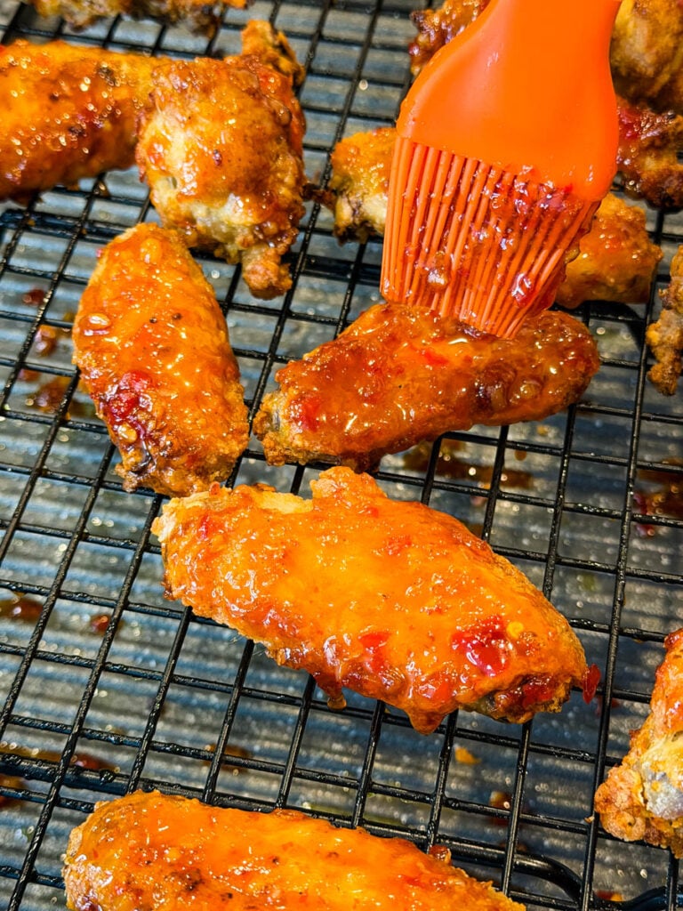 Crispy chicken wings being brushed with glossy sweet chili sauce while resting on a wire rack.