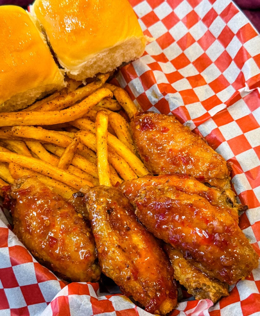 Close-up of crispy chicken wings coated in a sticky, glossy sweet chili sauce with seasoned fries and red and white checkered paper in the background.