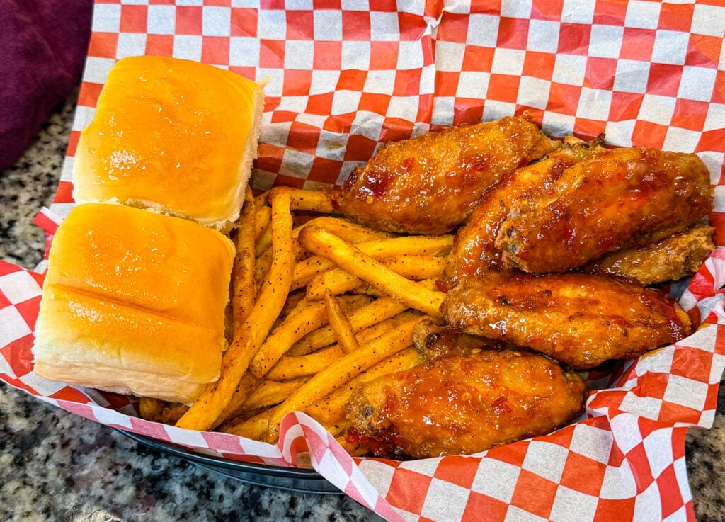 Basket of crispy sweet chili chicken wings coated in a glossy sauce and served with seasoned fries and buttery dinner rolls on red and white checkered paper.