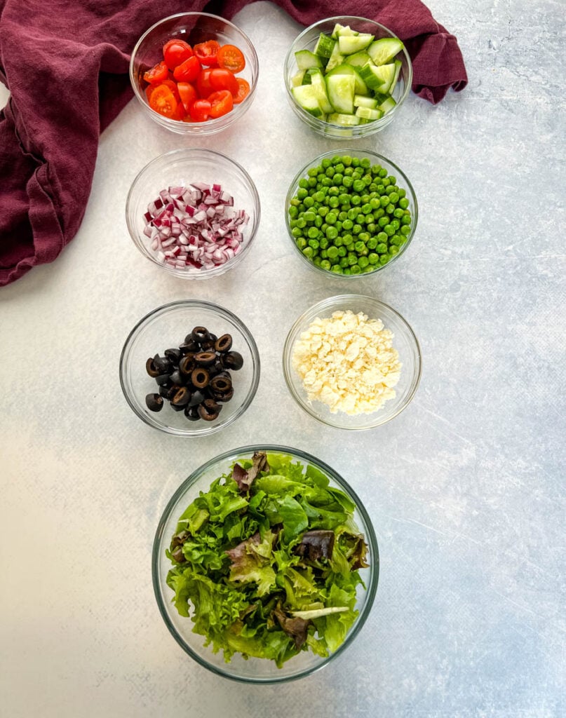 tomatoes, cucumbers, onions, peas, olives, feta, and fresh mixed greens in separate bowls