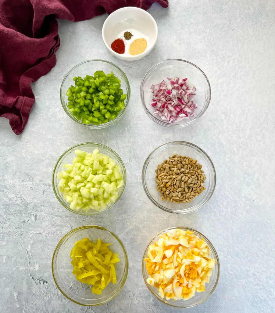 cucumbers, green peppers, red onions, sunflower seed kernels, pepperoncini, boiled eggs, and spices in separate glass bowls