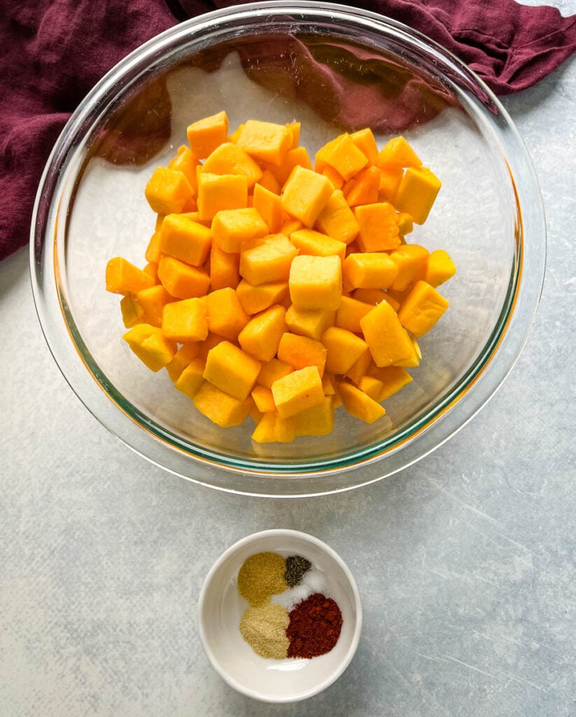 cut up butternut squash in a glass bowl along with a bowl of spices