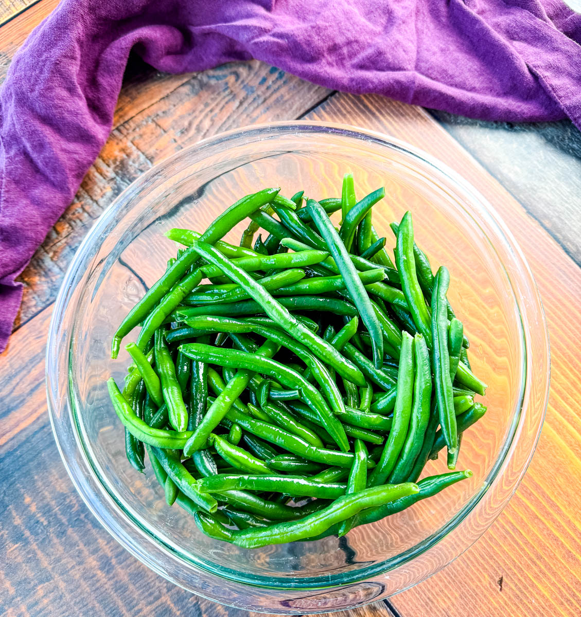 A clear glass bowl filled with bright green cooked green beans on a rustic wooden table with a purple napkin — fresh and ready for Southern green bean casserole.