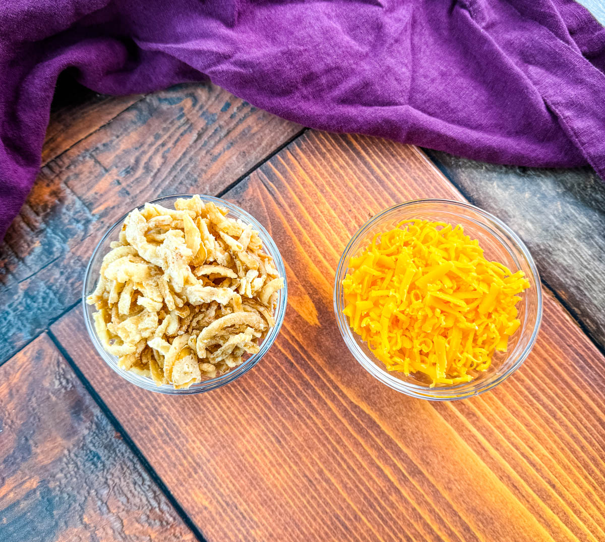 A small glass bowl of crispy fried onions and a bowl of shredded cheddar cheese sit on a wooden surface with a deep-purple napkin in the background — ingredients for Southern-style green bean casserole.