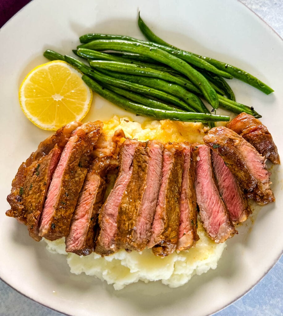 sliced lemon pepper steak with mashed potatoes and green beans on a white plate