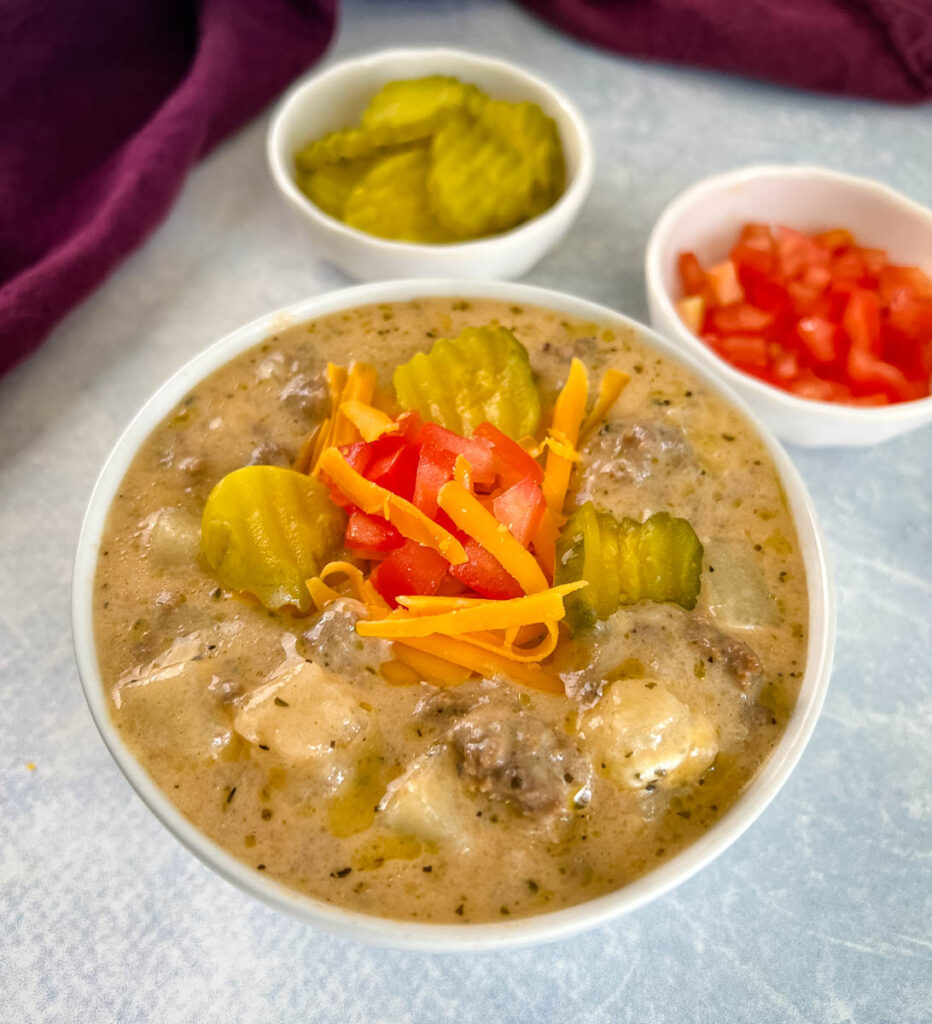 slow cooker cheeseburger soup in a white bowl with shredded cheese, pickles, and tomatoes