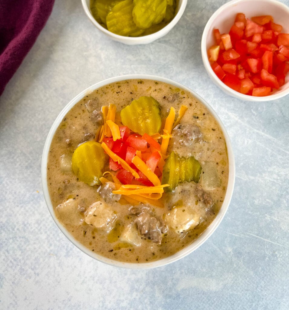 slow cooker cheeseburger soup in a white bowl with shredded cheese, pickles, and tomatoes