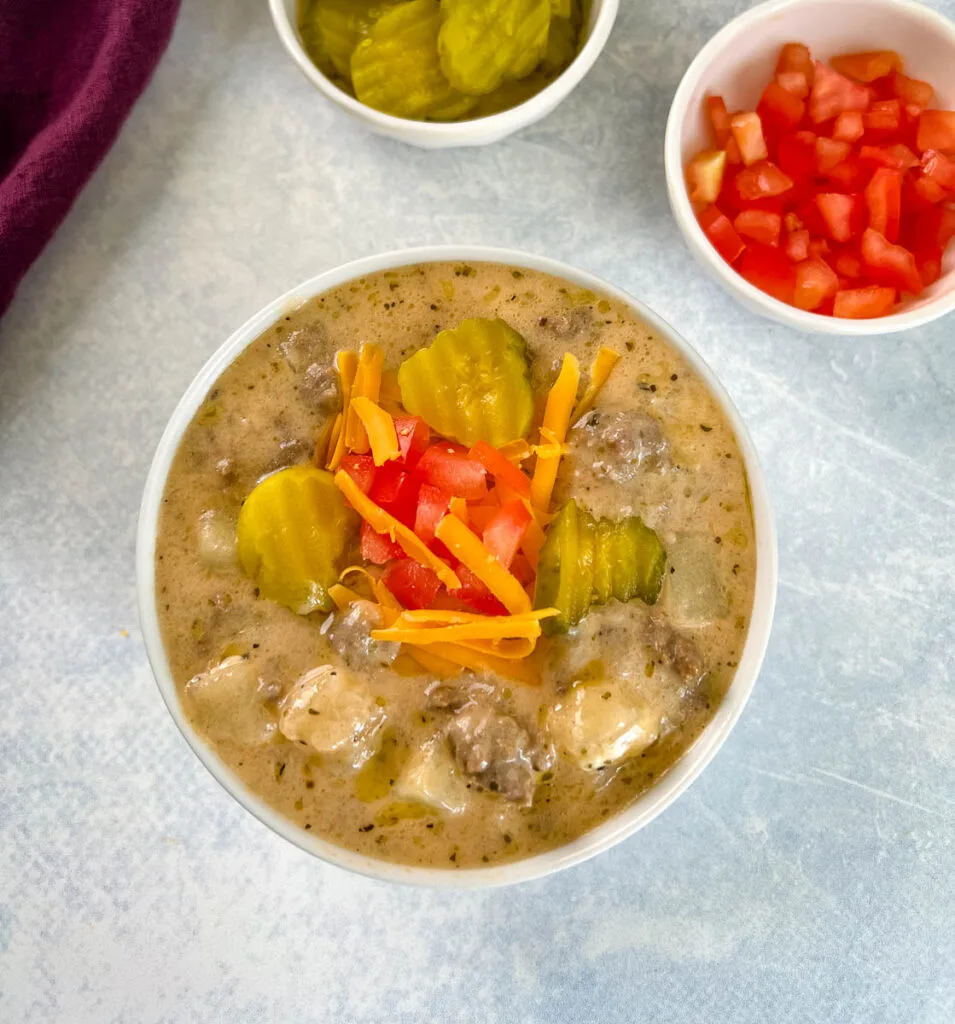 slow cooker cheeseburger soup in a white bowl with shredded cheese, pickles, and tomatoes