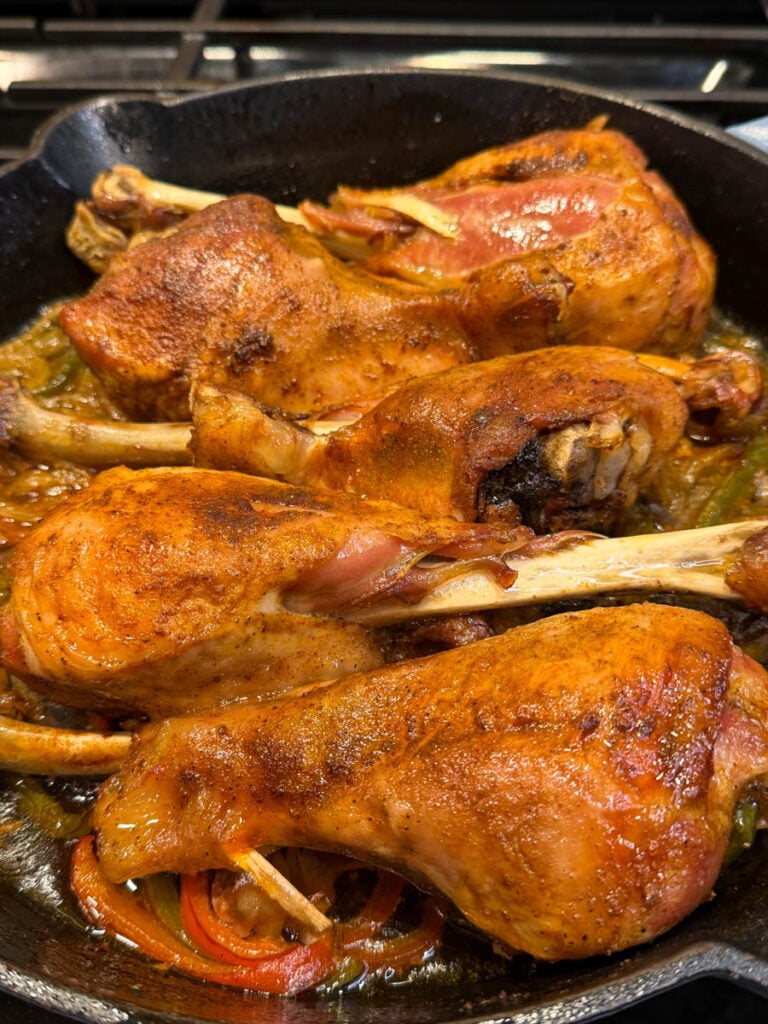 Close-up of baked turkey legs in a cast iron skillet, showing the juicy, seasoned skin and roasted vegetables beneath. The turkey looks tender and fall-off-the-bone, with visible steam and shine from the cooking juices.