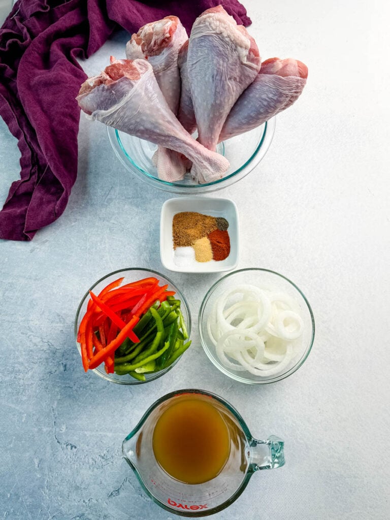 Raw turkey legs in a glass bowl surrounded by bowls of sliced red and green bell peppers, onions, a small dish of seasoning spices, and a measuring cup of broth. Everything is arranged neatly on a light surface with a purple linen in the corner.