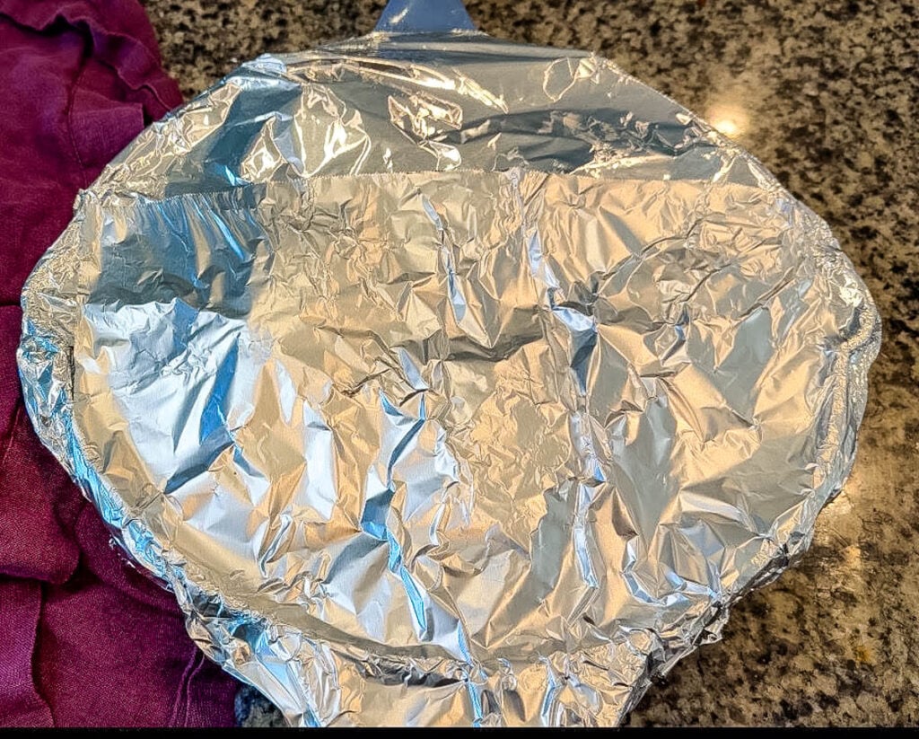 A baking dish covered tightly with aluminum foil, ready for the oven. A purple kitchen towel rests beside it on the countertop.
