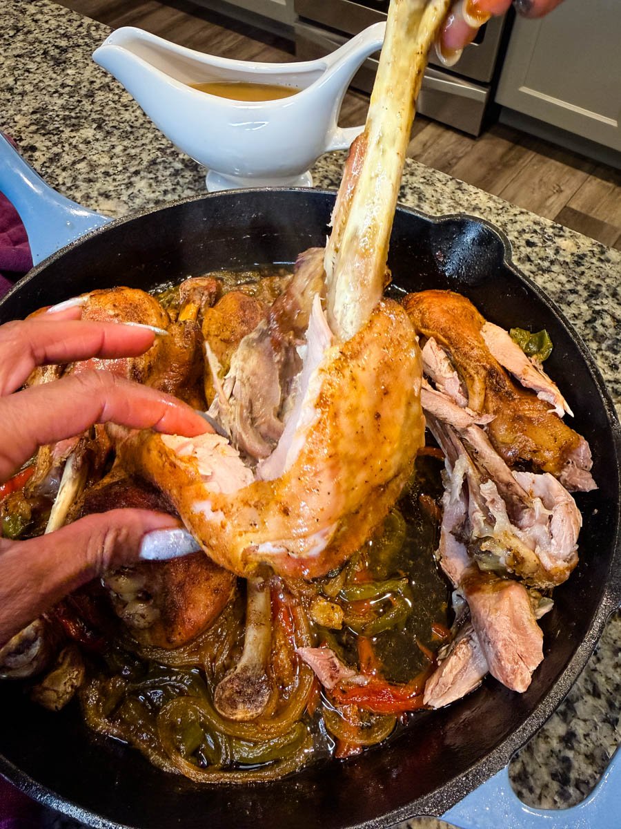 Hands pulling apart a tender baked turkey leg in a cast iron skillet filled with caramelized onions, bell peppers, and rich broth. The meat is visibly falling off the bone, showing how juicy and tender it is, with a gravy boat in the background.