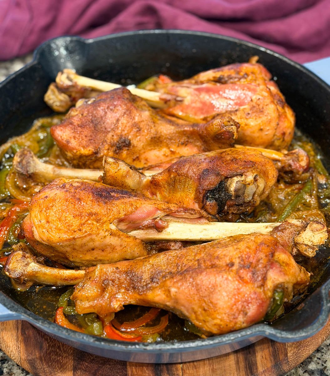 Golden brown baked turkey legs resting in a cast iron skillet with peppers and onions underneath. The turkey skin looks crisp and well-seasoned, with steam rising and juices bubbling at the bottom of the pan.