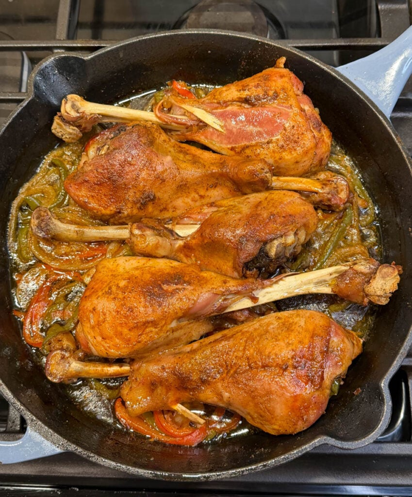 Four baked turkey legs resting in a cast iron skillet on the stove, surrounded by tender onions and bell peppers in a flavorful broth. The turkey skin is perfectly browned and glistening.