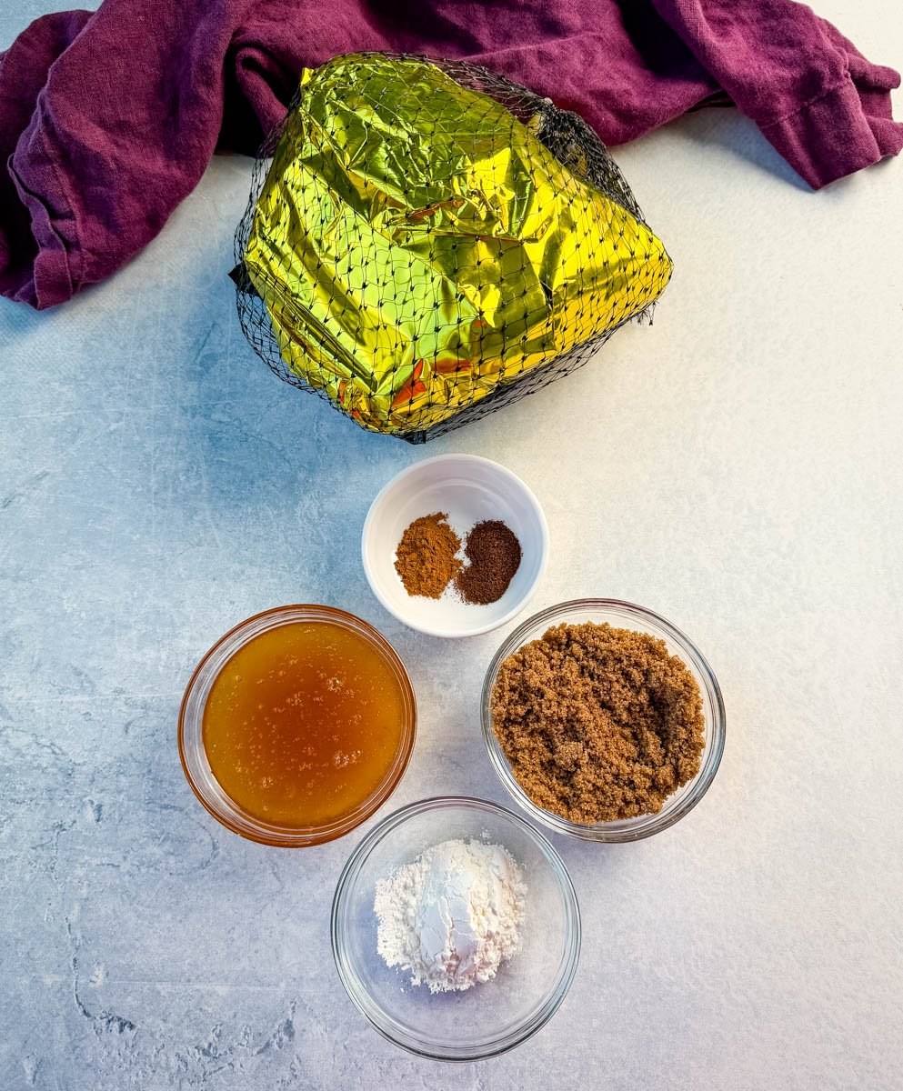 Ingredients laid out on a countertop next to a packaged ham wrapped in gold foil and netting. The ingredients include bowls of brown sugar, honey, cornstarch, and a small dish of warm spices like cinnamon and nutmeg.