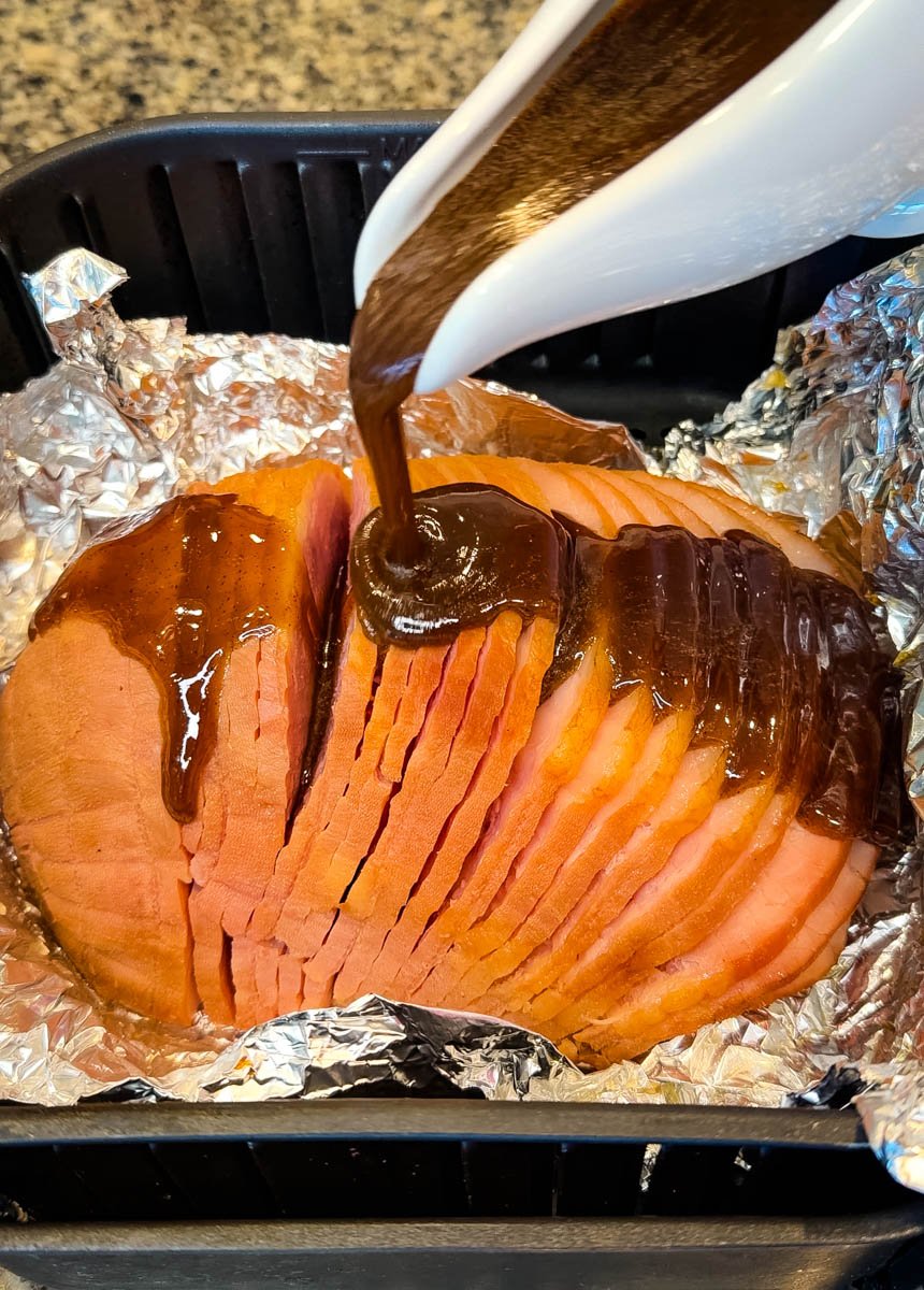 Thick brown glaze being poured over a sliced ham sitting on foil inside an air fryer basket, coating the ham for caramelization.