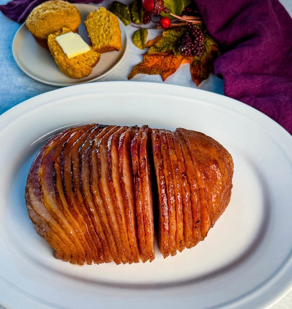 A full view of the spiral ham on a white serving platter, surrounded by a cozy setup with cornbread muffins, butter, and fall-themed decor in the background.