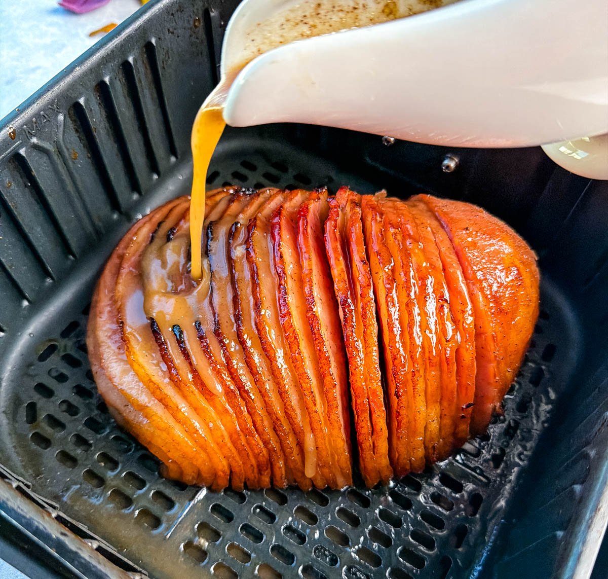 Honey glaze being poured from a white pitcher over a spiral ham inside the air fryer basket, coating it evenly for a glossy finish.