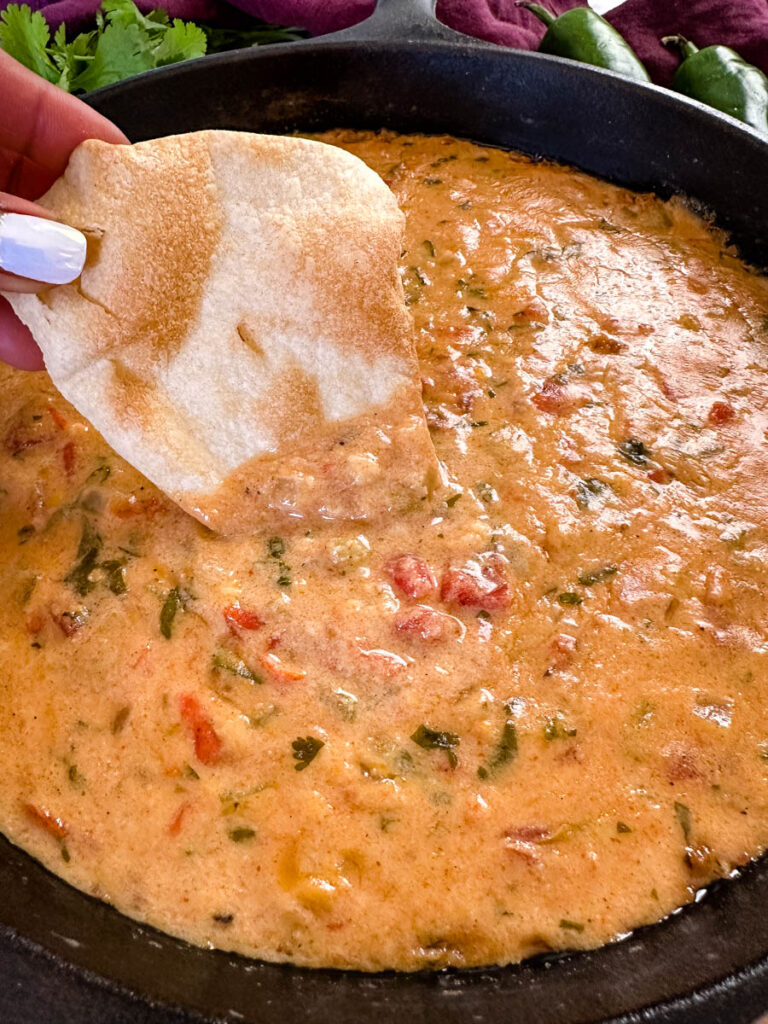 a person holding a chip dipped in smoked queso cheese dip in a cast iron skillet