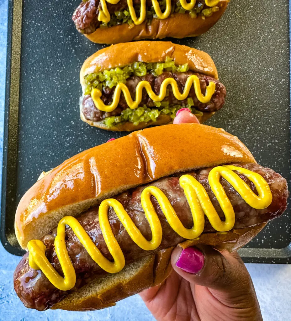 person holding smoked brat on buns with mustard and relish
