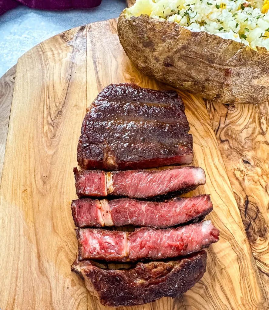 sliced smoked ribeye steak on a cutting board with a smoked baked potato