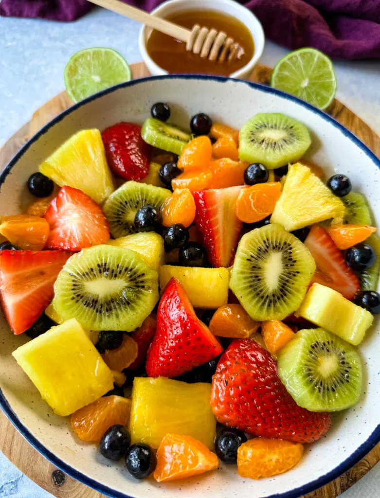 fresh strawberries, mandarin oranges, kiwi, blueberries, and pineapple fruit salad in a white bowl with honey lime dressing