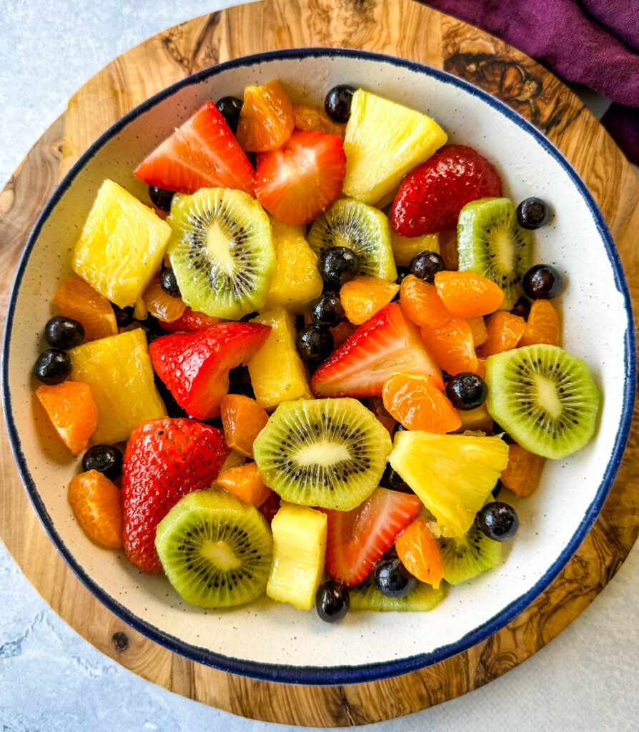 fresh strawberries, mandarin oranges, kiwi, blueberries, and pineapple fruit salad in a white bowl with honey lime dressing