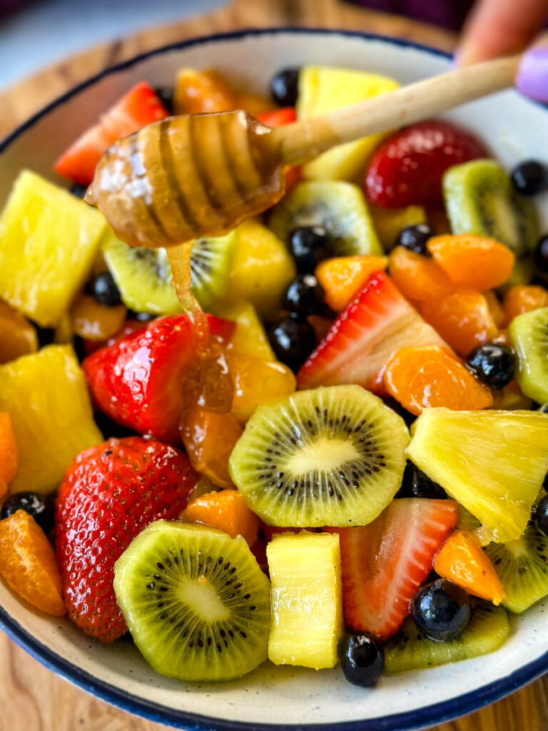 fresh strawberries, mandarin oranges, kiwi, blueberries, and pineapple fruit salad in a white bowl with honey lime dressing