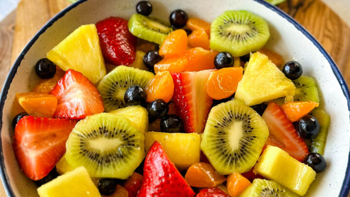 fresh strawberries, mandarin oranges, kiwi, blueberries, and pineapple fruit salad in a white bowl with honey lime dressing