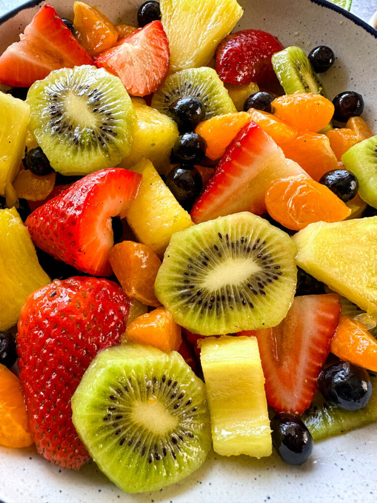 fresh strawberries, mandarin oranges, kiwi, blueberries, and pineapple fruit salad in a white bowl with honey lime dressing