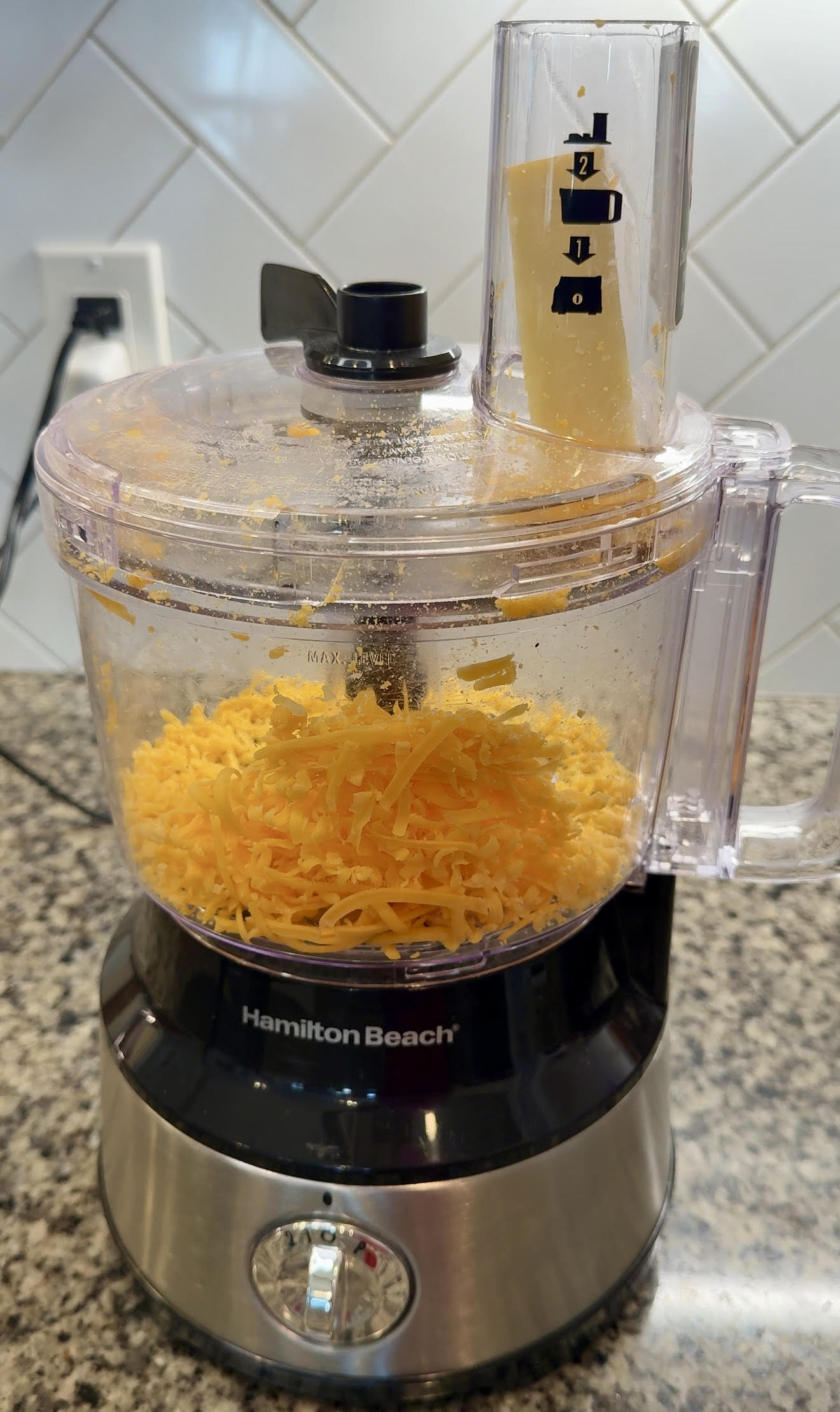 Food processor with a block of cheddar cheese in the chute, ready to be shredded.