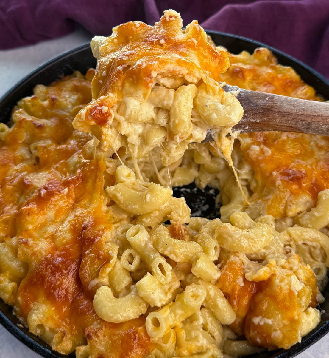 Close-up of a cast iron skillet filled with Southern baked mac and cheese, with a wooden spoon lifting a gooey serving showing melted cheddar cheese stretching.