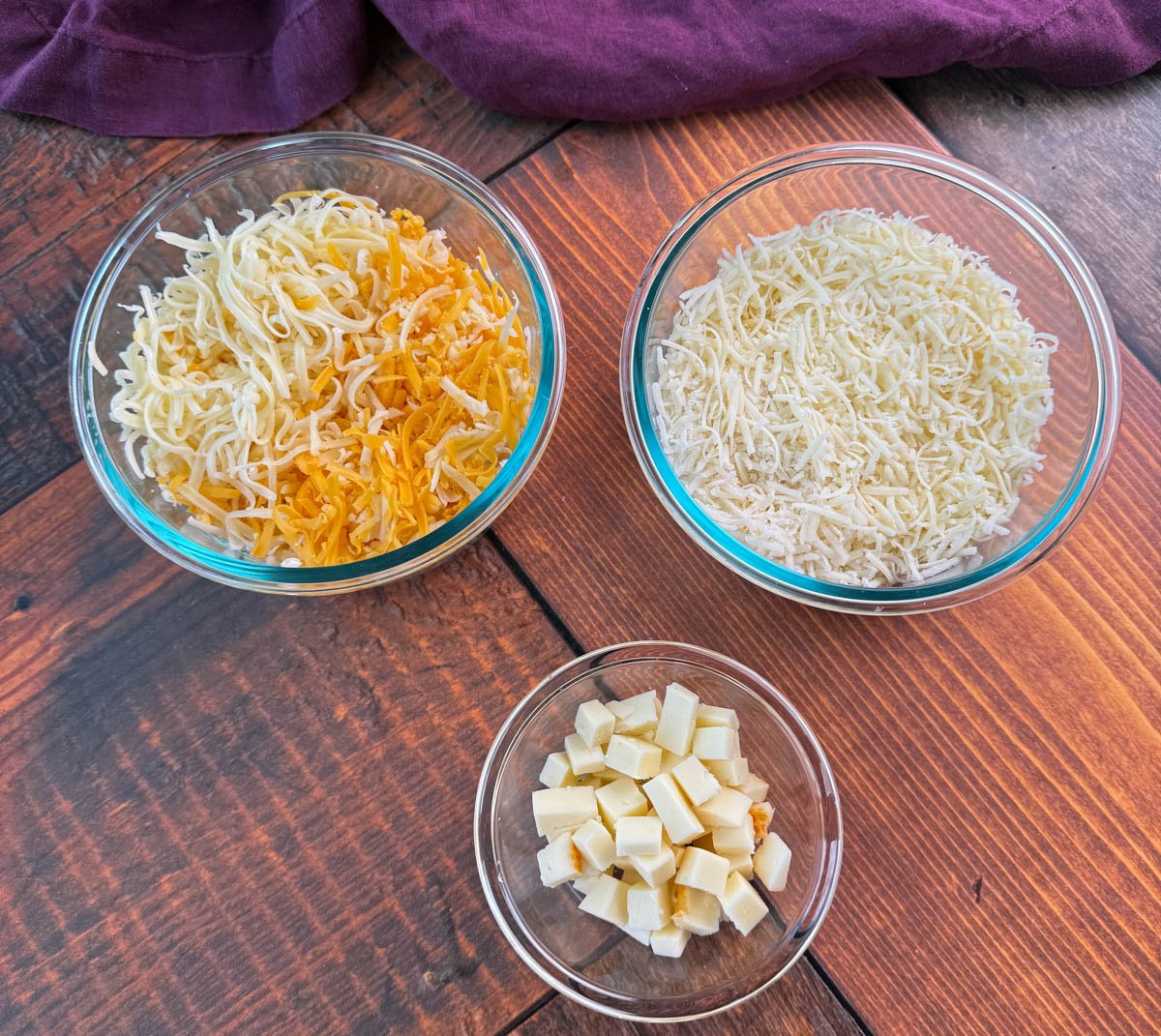 Overhead view of three glass bowls on a wooden table—one with shredded cheddar and mozzarella, one with shredded white cheese, and one small bowl with cubed cheese.
