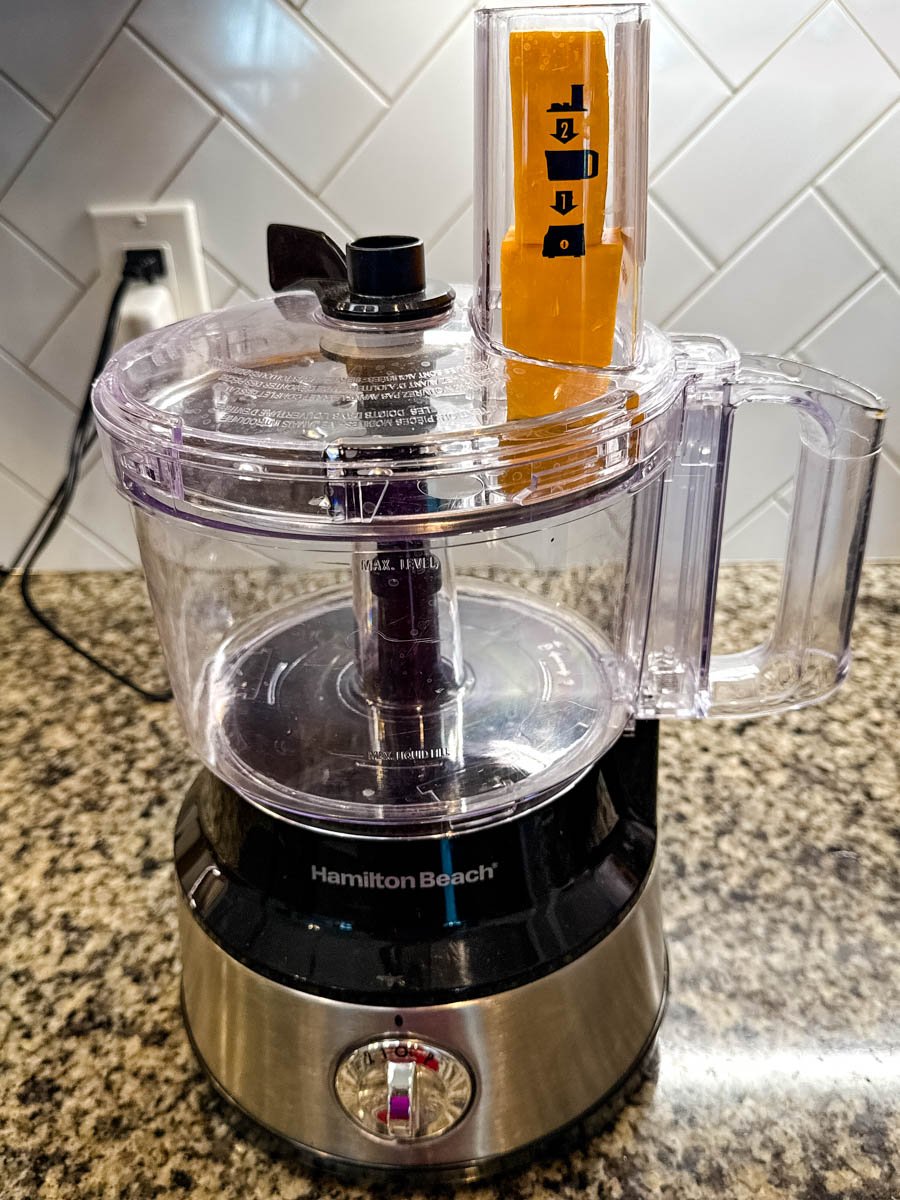 Food processor with a block of cheddar cheese in the chute, ready to be shredded.