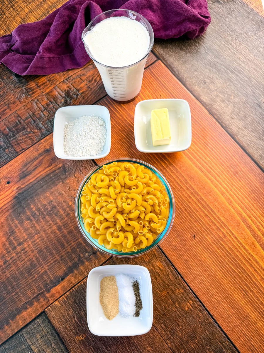 Overhead photo of mac and cheese ingredients on a wooden surface, including elbow macaroni, flour, butter, milk, and small bowls of garlic powder, salt, and pepper.