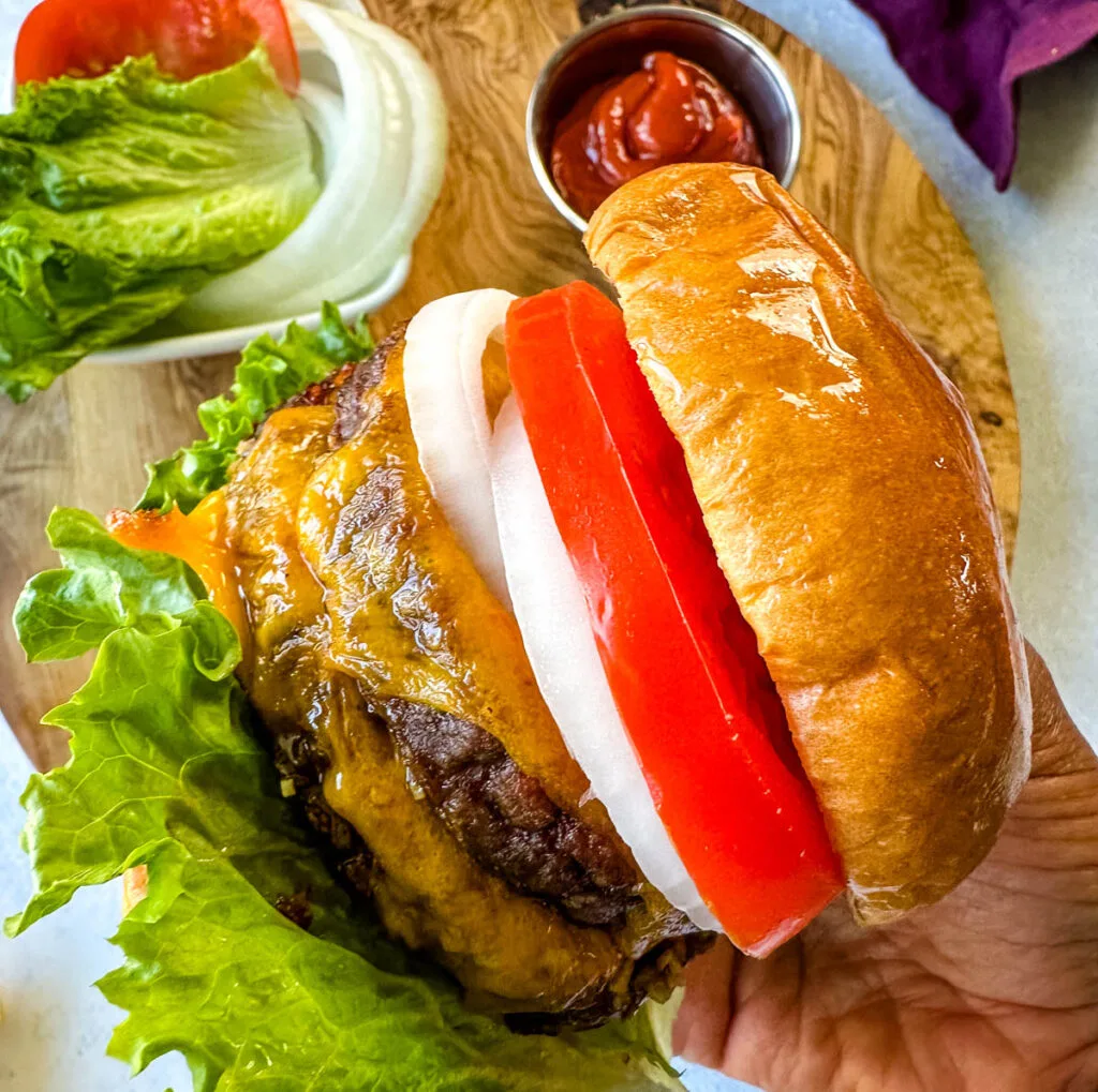 person holding Traeger smoked burger on a bun with cheese, lettuce, tomato, and onions