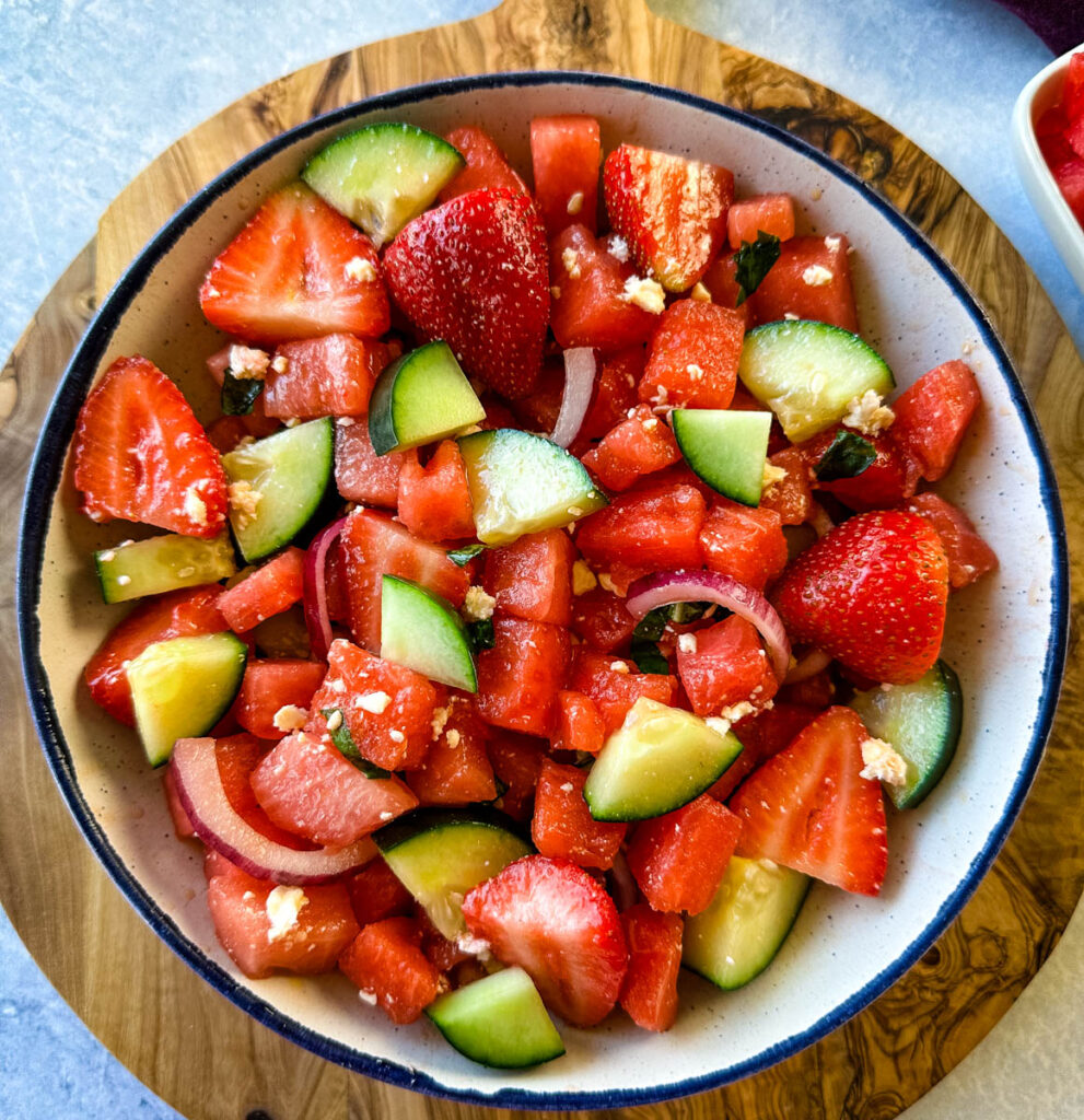 watermelon salad with feta, basil, cucumbers, avocado, strawberries, onion, and balsamic lime dressing in a white bowl