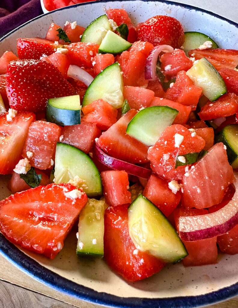 watermelon salad with feta, basil, cucumbers, avocado, strawberries, onion, and balsamic lime dressing in a white bowl