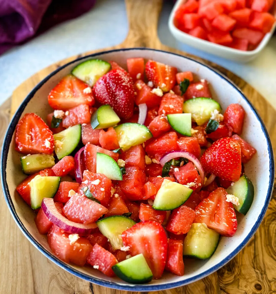 watermelon salad with feta, basil, cucumbers, avocado, strawberries, onion, and balsamic lime dressing in a white bowl