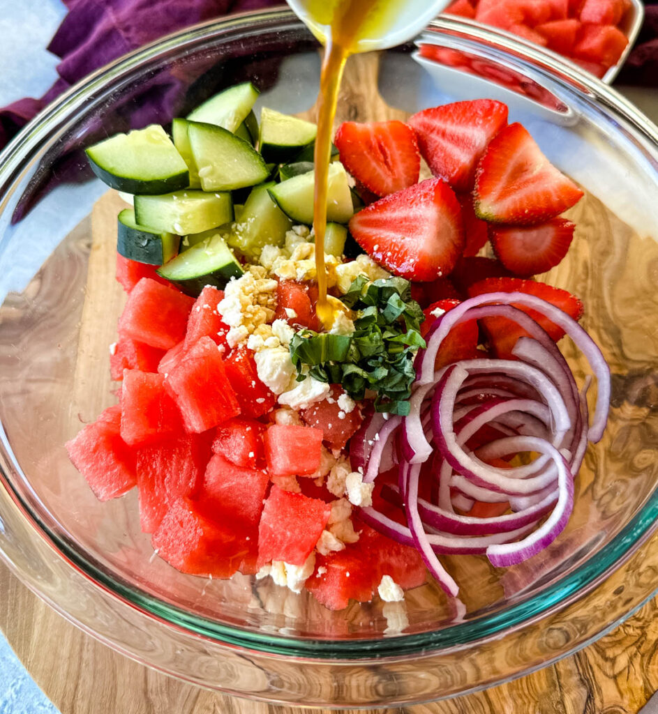 watermelon salad with feta, cucumbers, avocado, basil, strawberries, onion, and balsamic lime dressing in a glass bowl drizzled with balsamic dressing