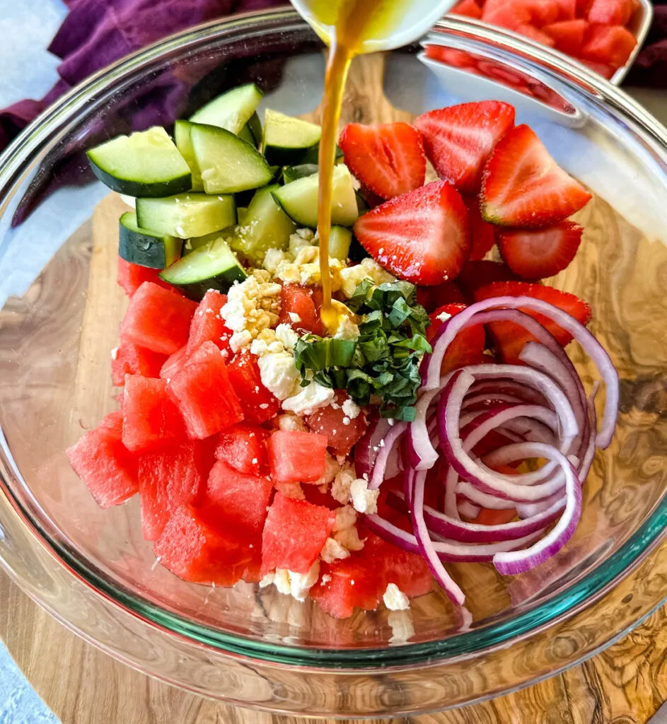 watermelon salad with feta, cucumbers, avocado, basil, strawberries, onion, and balsamic lime dressing in a glass bowl drizzled with balsamic dressing
