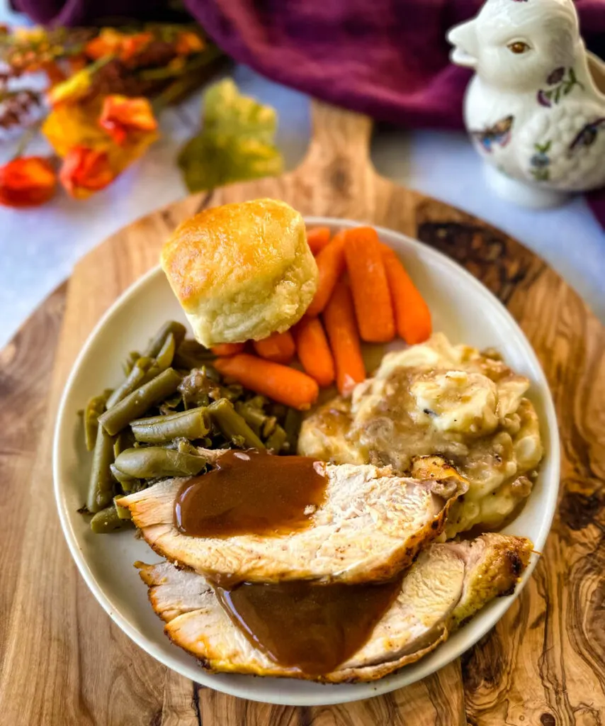 sliced bone in turkey breast on a plate with gravy, mashed potatoes, green beans, carrots, and biscuits