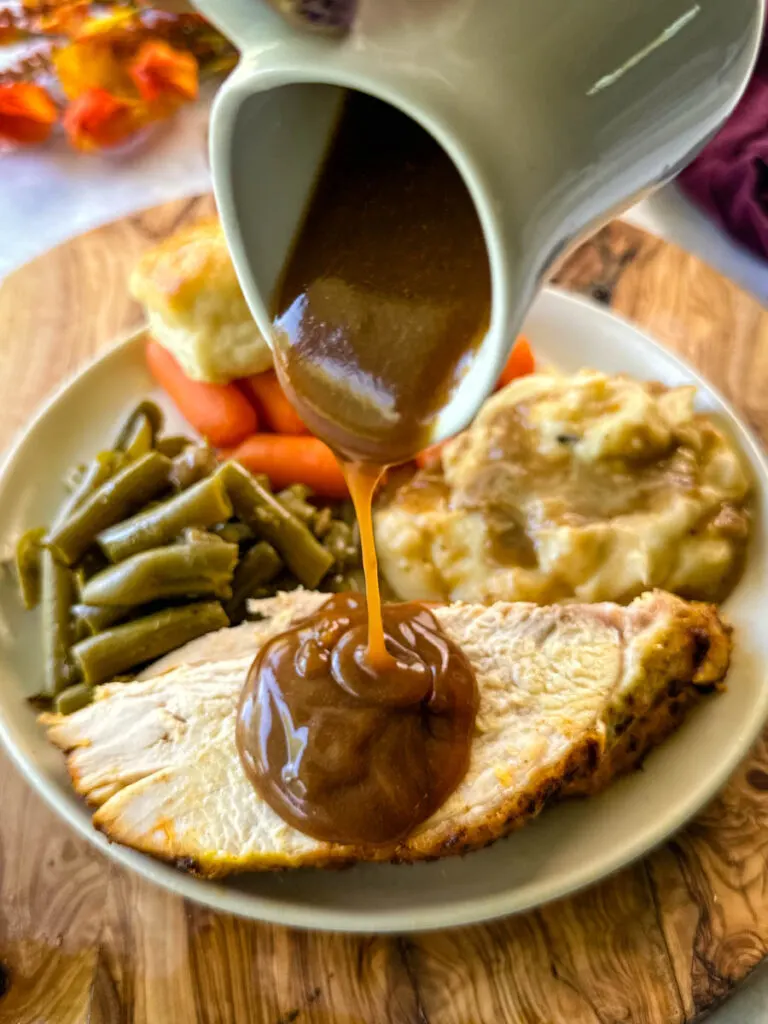 gravy drizzled over sliced bone in turkey breast on a plate with mashed potatoes, green beans, carrots, and biscuits