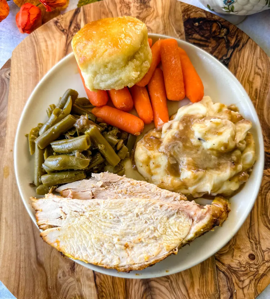 sliced bone in turkey breast on a plate with gravy, mashed potatoes, green beans, carrots, and biscuits
