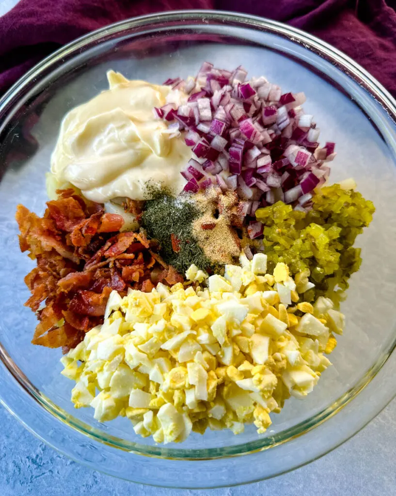 bacon pieces, onions, relish, diced hard boiled eggs, and ranch seasoning over potatoes in a glass bowl