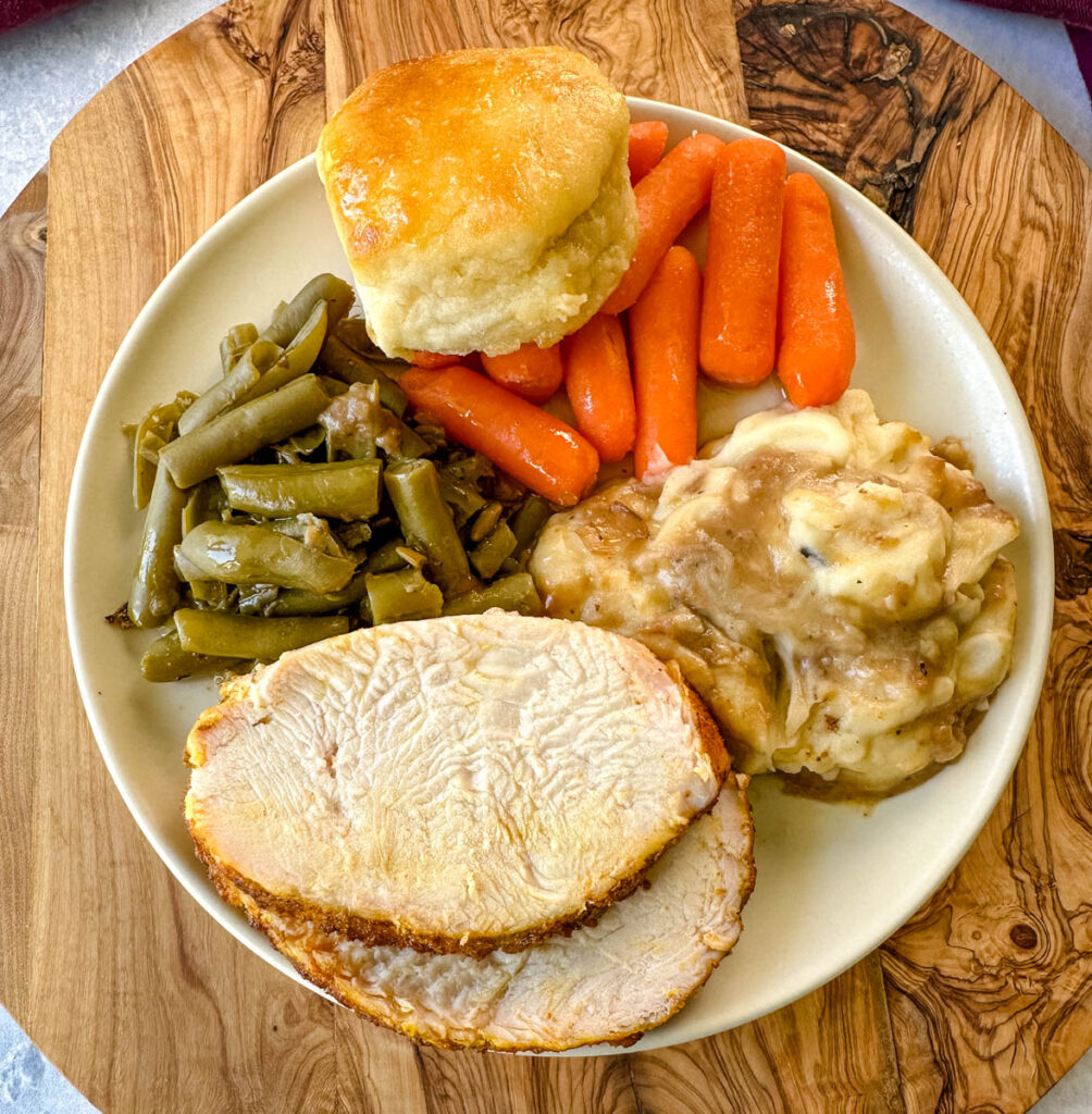boneless turkey breast slices on a plate with mashed potatoes, gravy, green beans, carrots, and a biscuit