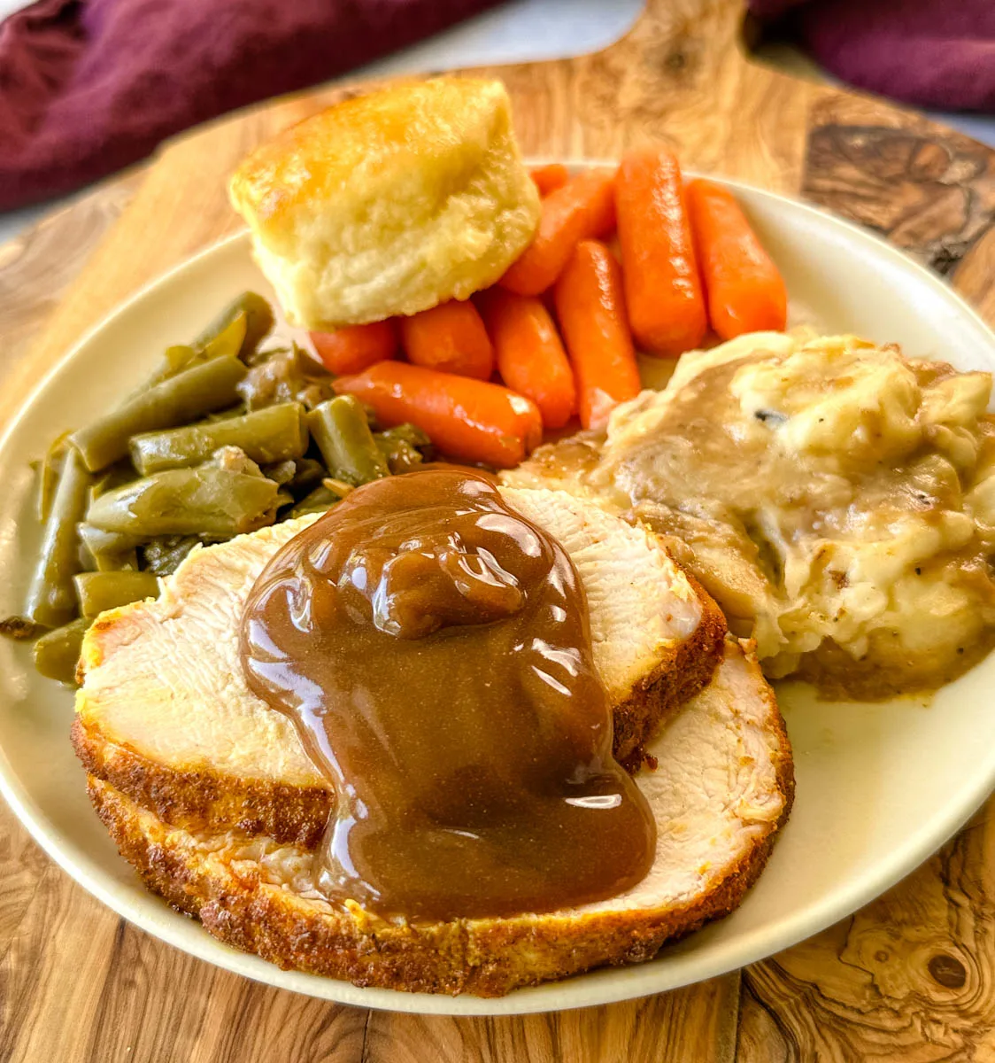 boneless turkey breast slices on a plate with mashed potatoes, gravy, green beans, carrots, and a biscuit