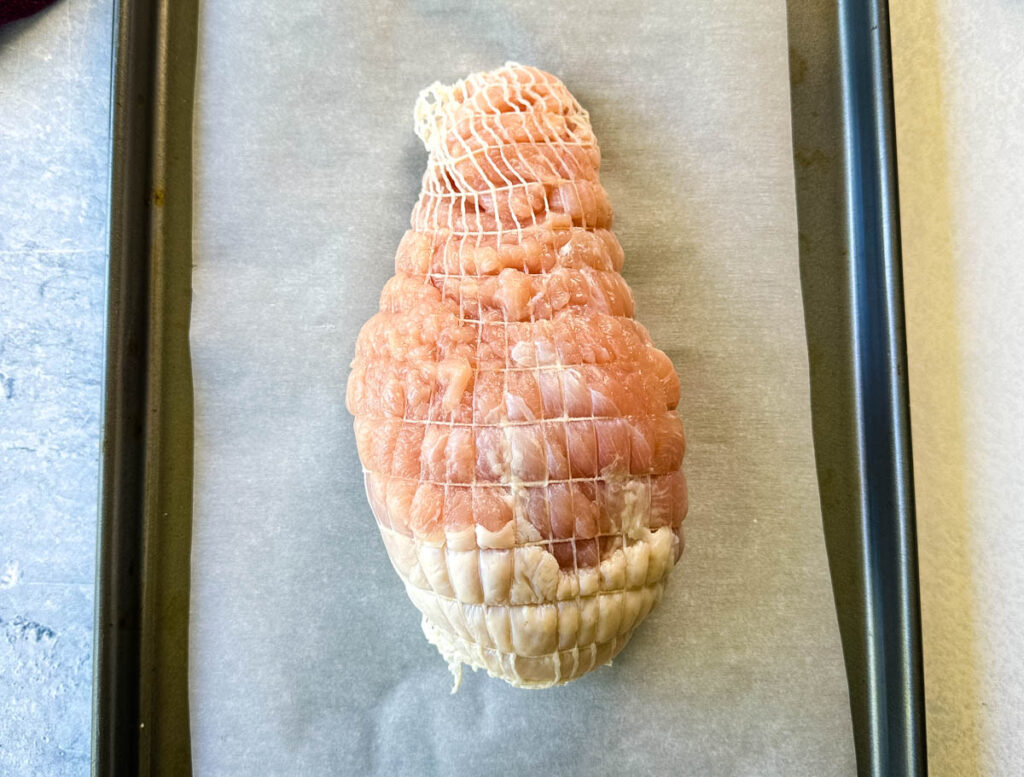 raw, seasoned boneless turkey breast wrapped in a net on a parchment paper lined sheet pan